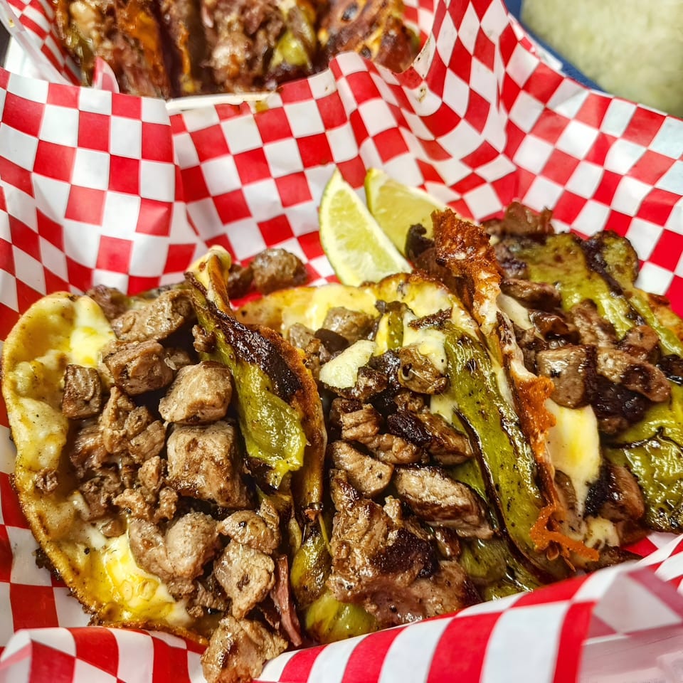 Three sizzling tacos with steak, melted cheese, and green chiles in a red and white checkered basket.