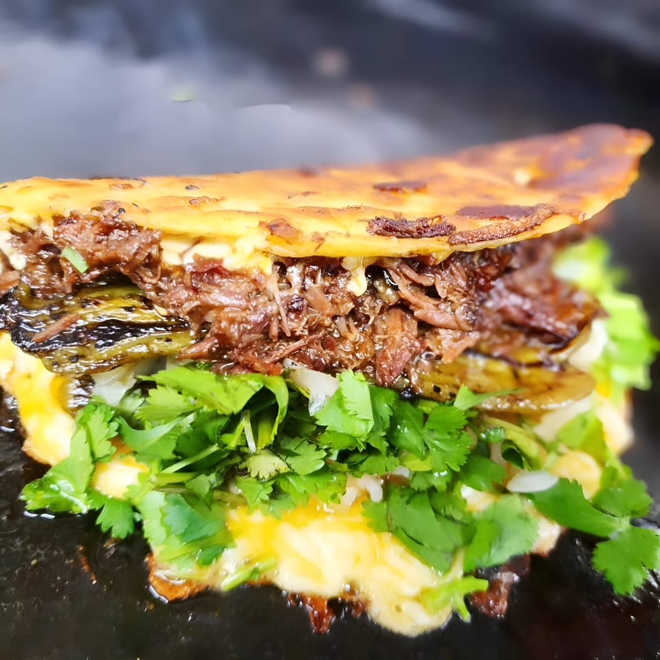 A close-up of a sizzling birria taco from The Taco Man, filled with meat, cheese, and cilantro.