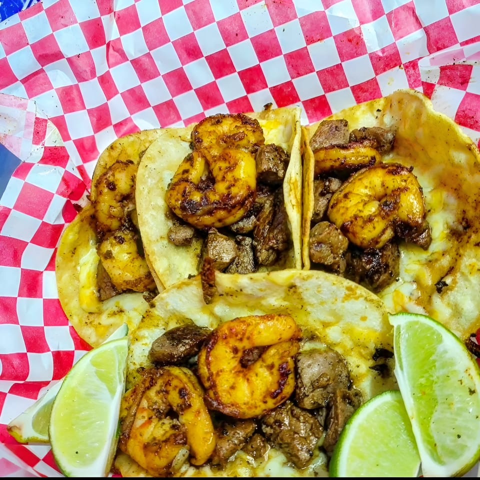Four shrimp and steak tacos with lime wedges, served in a red and white checkered basket.