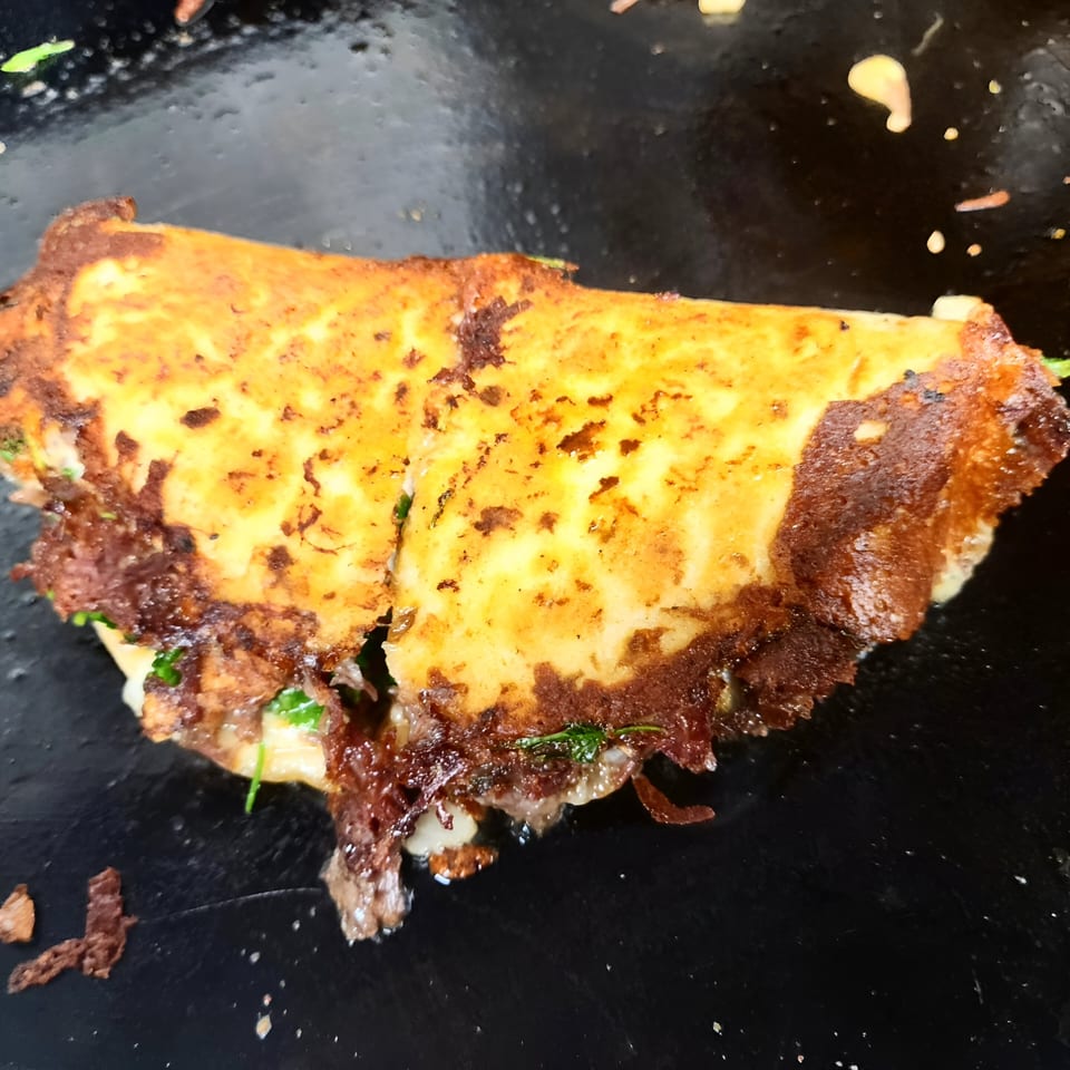 A golden-brown, crispy quesadilla from The Taco Man, filled with meat and cilantro, on a dark surface.