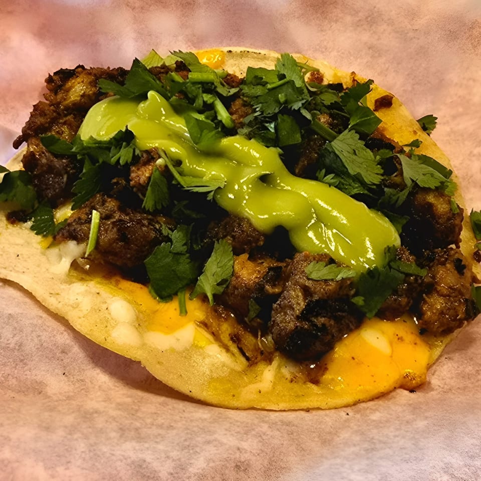 A close-up of a The Taco Man taco with meat, cilantro, and green sauce on a corn tortilla.